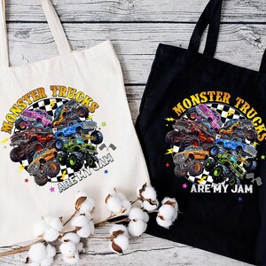 two tote bags with a vibrant graphic design of monster trucks. The bags are placed on a wooden surface, with a branch of cotton balls in the foreground.