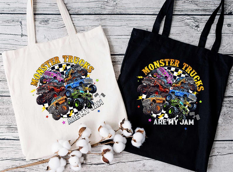 two tote bags with a vibrant graphic design of monster trucks. The bags are placed on a wooden surface, with a branch of cotton balls in the foreground.