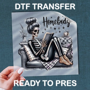May include: A DTF transfer sheet featuring a skeleton with a high bun hairstyle sitting on a tufted sofa reading a book. The text reads DTF TRANSFER, Homebody, and READY TO PRES. The skeleton wears leopard print socks and sits near a small plant.