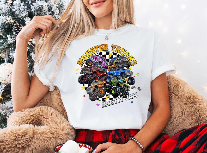 A woman wearing a white t-shirt with a graphic of monster trucks and the text "MonSTER TRUCKS ARE MY JAM" is sitting in front of a Christmas tree.