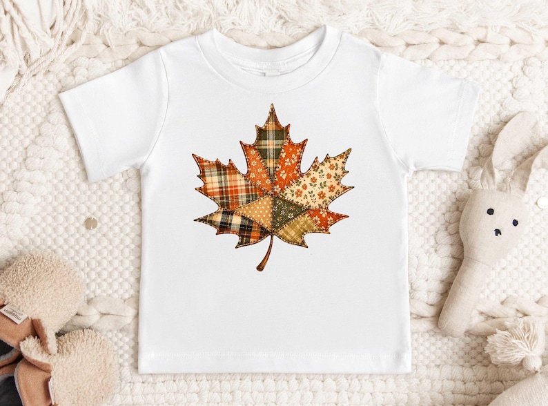 A white t-shirt with a colorful patchwork leaf design printed on it, placed on a white surface with a stuffed rabbit and other plush toys nearby.