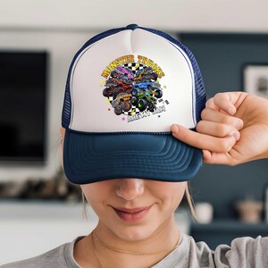 A person is holding up a white and blue baseball cap with a colorful graphic of monster trucks on it.