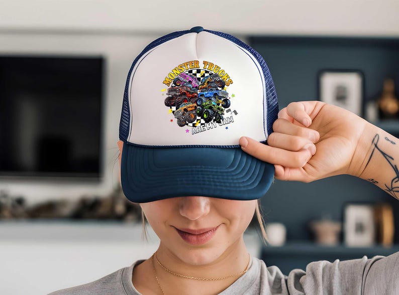 A person is holding up a white and blue baseball cap with a colorful graphic of monster trucks on it.