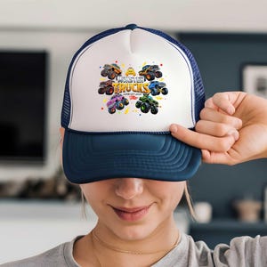 A person is holding up a white and blue baseball cap with a colorful graphic of monster trucks on it.