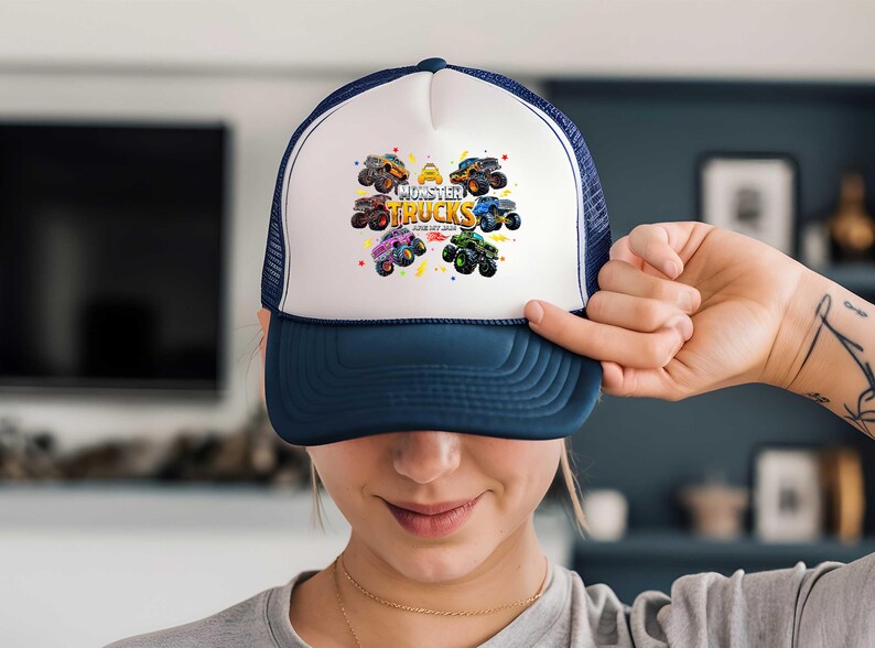 A person is holding up a white and blue baseball cap with a colorful graphic of monster trucks on it.