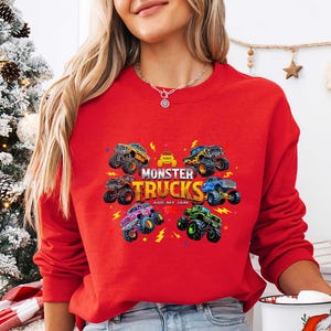 A woman wearing a red sweater with a graphic of monster trucks and the text "MONSTER TRUCKS ARE MY LIGHT" is standing in front of a Christmas tree.