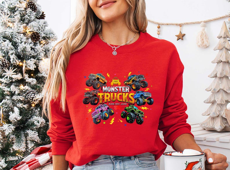 A woman wearing a red sweater with a graphic of monster trucks and the text "MONSTER TRUCKS ARE MY LIGHT" is standing in front of a Christmas tree.