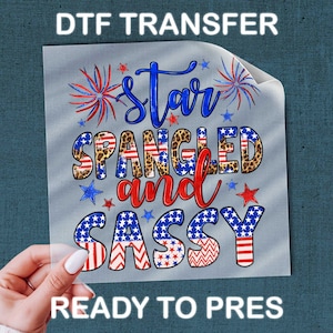 May include: A DTF transfer with the text "Star Spangled and Sassy" in red, blue, and white, with star and firework graphics. The transfer is held by a hand with white nail polish.