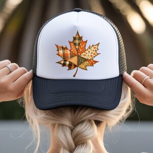 A person is adjusting a white baseball cap with a black brim featuring a colorful autumn leaf design.