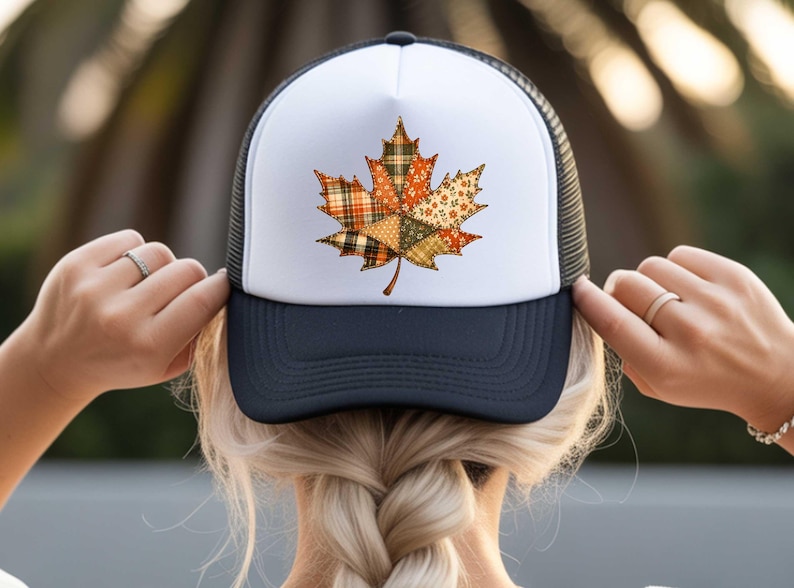 A person is adjusting a white baseball cap with a black brim featuring a colorful autumn leaf design.