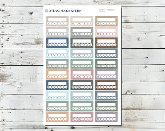 May include: A sheet of planner stickers with the text "MTWTFSS" in various colors, including blue, green, and brown. The stickers are rectangular with a white space for writing. The top of the sheet has the text "ATLAS DESIGN STUDIO" and "Habits/Neutrals".