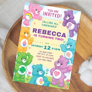 以下が含まれることがあります： ケアベアのキャラクターが描かれたカラフルな招待状。「ご招待！」と「レベッカは2歳になります！」という文字とパーティーの日時が記載されています。招待状は木製のトレイの上にあります。