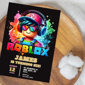 May include: A black invitation card featuring the Roblox logo and a cartoon character wearing headphones and sunglasses. The card reads "JAMES IS TURNING SIX!" with event details. The card is on a wooden plate.