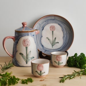 Peut inclure: Un service à thé en céramique comprenant une théière bleue et blanche, une assiette et deux tasses. Chaque pièce est ornée d'un motif de tulipe rose. L'ensemble est disposé sur une surface en bois avec des brins d'herbes.