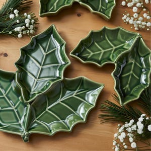 Op de afbeelding: Een verzameling glanzende groene keramische schalen in de vorm van bladeren. De schalen hebben gedetailleerde nerven en zijn gerangschikt op een houten oppervlak, met takjes groen en witte bloemen.