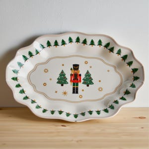 May include: A white, scalloped-edge serving platter with a Christmas theme. The platter features a central design of a nutcracker soldier and two green Christmas trees. The border is decorated with green Christmas trees.