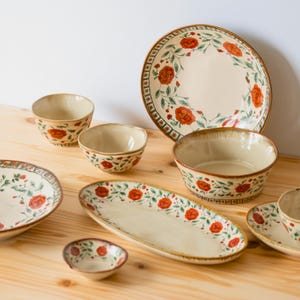 May include: A selection of cream-coloured ceramic tableware with a floral design of red flowers and green leaves. The set includes bowls, plates, and a serving platter, all with a rustic, hand-painted aesthetic. The rims have a brown border.