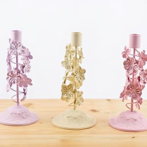 May include: Three decorative candle holders in pastel colors. The holders feature floral designs and are painted in shades of lavender, cream, and pink. The candle holders are made of metal and sit on a wooden surface.