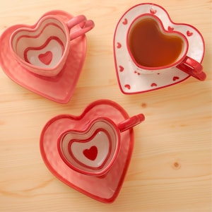 Puede incluir: Tres tazas de té y platillos en forma de corazón en tonos rosa y blanco. Una taza está llena de té. Las tazas y los platillos tienen un diseño de corazón. El juego está sobre una superficie de madera clara.