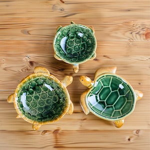 Peut inclure: Trois bols en céramique en forme de tortue avec glaçure verte et jaune. Les bols présentent des motifs de carapace détaillés et sont disposés sur une surface en bois. Les bols tortues sont décoratifs et fonctionnels.