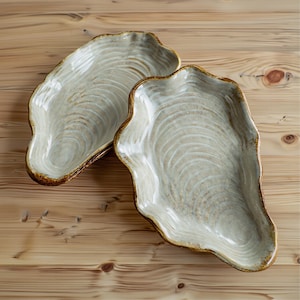 May include: Two ceramic serving platters, oyster shell-shaped, in light beige with concentric wave patterns and a dark brown rim. The background is a light brown wooden surface. Each platter is approximately 30 cm in length.