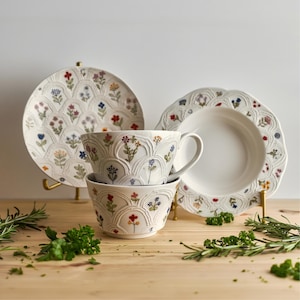 Può includere: Un set di stoviglie in ceramica bianca con un motivo floreale. Il set comprende un piatto grande, una ciotola e due tazze. Il motivo floreale presenta fiori rossi, blu e gialli. Il set è esposto su una superficie di legno.