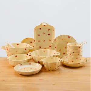 Può includere: Una collezione di stoviglie in ceramica color crema con un motivo a cuore. Il set include ciotole, piatti e un piatto da portata. Il motivo a cuore è di colore rosso-arancio. Le stoviglie sono disposte su una superficie di legno.