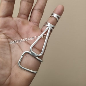 以下が含まれることがあります： シルバーの指輪セット。 セットには、指の先端用のリング、中央用のリング、指を下る接続部分が含まれています。 テキスト「AMBERANDAMETHYSTT」が表示されています。