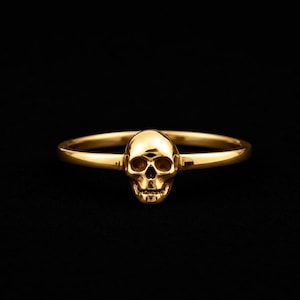 Puede incluir: Un anillo dorado con un diseño detallado de calavera. La calavera es el punto focal, con cuencas de los ojos, cavidad nasal y dientes. La banda del anillo es delgada y lisa, creando una estética minimalista pero audaz. El anillo está sobre un fondo negro.