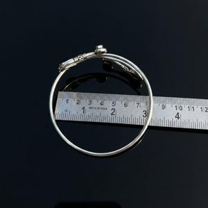 May include: A silver-toned metal bangle bracelet featuring a bypass design with decorative wire-wrapped ends. A metal ruler is positioned across the center of the bracelet, showing an internal diameter of approximately 2.5 inches.