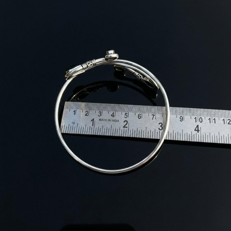 May include: A silver-toned metal bangle bracelet featuring a bypass design with decorative wire-wrapped ends. A metal ruler is positioned across the center of the bracelet, showing an internal diameter of approximately 2.5 inches.