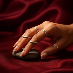 May include: A hand wearing several thin, silver rings. The rings are simple bands, and the hand is resting on a small, smooth, gray stone. The background is a deep red, silky fabric.