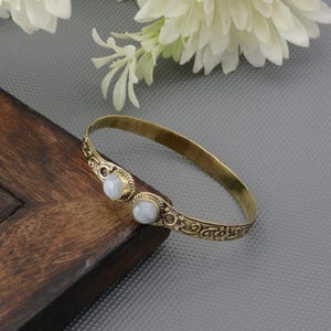 May include: A brass-toned bracelet with an open design. The bracelet features two round, light-colored stones set in decorative bezels. Intricate scrollwork adorns the bracelet's surface, adding a vintage aesthetic. The bracelet is displayed on a wooden surface.