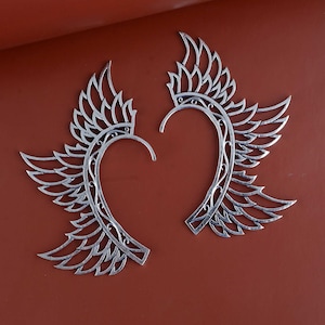 May include: Two silver-colored ear cuffs, each shaped like a stylized wing. The wings have intricate feather detailing and a curved design that wraps around the ear. The background is a solid, warm brown.