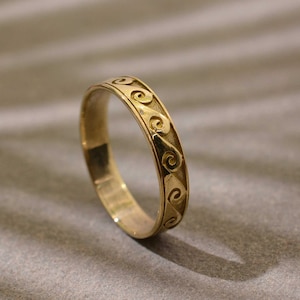 Puede incluir: Un anillo dorado con un patrón de olas repetido grabado alrededor de la banda. El anillo se presenta sobre un fondo gris neutro, resaltando el intrincado diseño y el cálido tono metálico. La superficie del anillo refleja la luz, mostrando su acabado pulido.