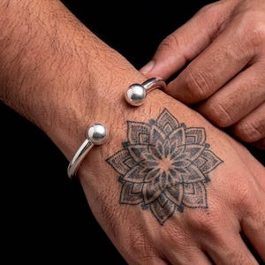 May include: A polished silver cuff bracelet featuring a smooth, open-ended design with two rounded ball terminals at each end. The bracelet is crafted from a solid, reflective metal band that curves around the wrist, showcasing a minimalist aesthetic.