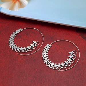 May include: A pair of silver hoop earrings. Each earring has a spiral design with a decorative, openwork crescent shape. The crescent is decorated with small circles and dots. The earrings are displayed on a red surface.
