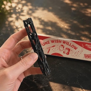 May include: A black, cylindrical, porous object with an irregular, hollowed-out lattice structure. Beside it sits a rectangular white and red box labelled ONE WISH WILLOW, featuring illustrations of two faces, the word CRAKK, and the text AMAZE YOUR FRIENDS!