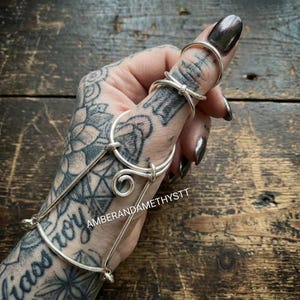 Può includere: Gioielli per le mani color argento con più anelli e un bracciale di collegamento. Gli anelli sono collegati da barre delicate e un vortice decorativo. I gioielli sono indossati su una mano tatuata. Il testo "AMBERANDAMETHYSTT" è visibile.
