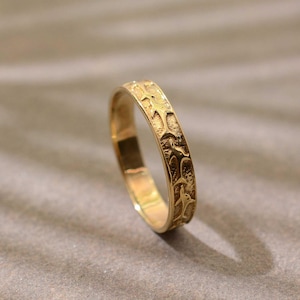 Peut inclure: Une bague dorée ornée d'un motif répétitif de formes étoilées stylisées. La surface de la bague est texturée et réfléchit la lumière. La bague est présentée sur un fond neutre. Le motif est gravé.