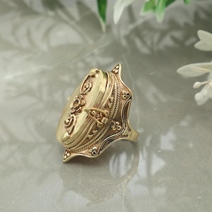 Könnte beinhalten: Ein goldfarbener Medaillonring mit einem verzierten Design. Der Ring hat ein rechteckiges Medaillon mit floralen und scrollartigen Details, einen kleinen Verschluss und ein aufwendig gestaltetes Band. Der Ring ist vor einem neutralen Hintergrund platziert.