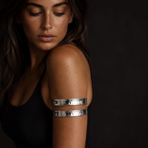 May include: Two silver arm cuffs with a hammered texture are worn on the upper arm. The jewelry is displayed against a dark background. The model is wearing a black tank top.