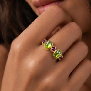 Peut inclure: Une bague en or ornée de deux cabochons ovales en péridot vert et de trois petites améthystes violettes de taille ronde, disposées en ligne sur l'anneau. Les pierres sont serties dans des montures en or jaune poli à la finition lisse.