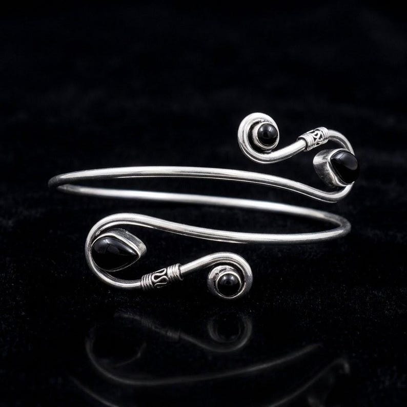 May include: A silver bypass bracelet featuring a spiral design at each end. Each terminal is accented with a smooth, polished black stone, including one teardrop-shaped stone and three round stones set within the metallic silver wire frame.