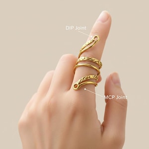 May include: A gold-coloured, multi-finger ring with a unique, coiled design. The ring wraps around two fingers, featuring textured detailing and open loops at the finger joints. The ring is worn on a hand.
