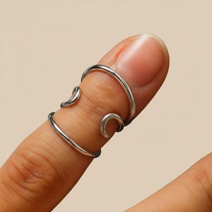 May include: A silver finger ring with a distinctive open design. The ring wraps around the finger with curved accents, creating an elegant and modern look. The ring is shown on a finger against a neutral background.