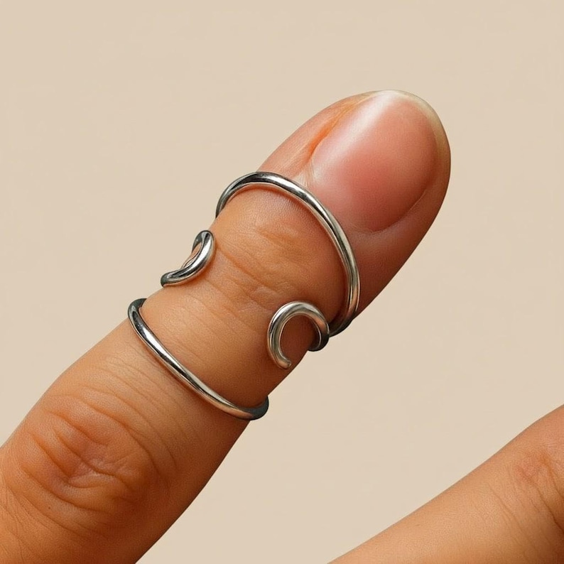 May include: A silver finger ring with a distinctive open design. The ring wraps around the finger with curved accents, creating an elegant and modern look. The ring is shown on a finger against a neutral background.