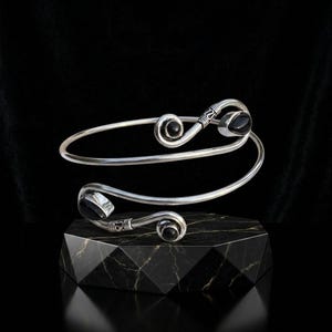 May include: A silver spiral cuff bracelet featuring two polished black stones at each end. The metal band has a smooth finish with decorative etched detailing near the terminals, creating a continuous wrap-around design that is open at the center.