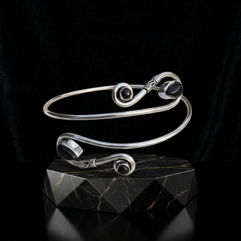 May include: A silver spiral cuff bracelet featuring two polished black stones at each end. The metal band has a smooth finish with decorative etched detailing near the terminals, creating a continuous wrap-around design that is open at the center.
