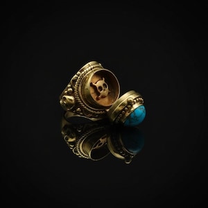May include: A gold-colored ring with an open compartment revealing a skull emblem. The ring features intricate detailing and a turquoise stone. The ring is displayed on a reflective black surface.